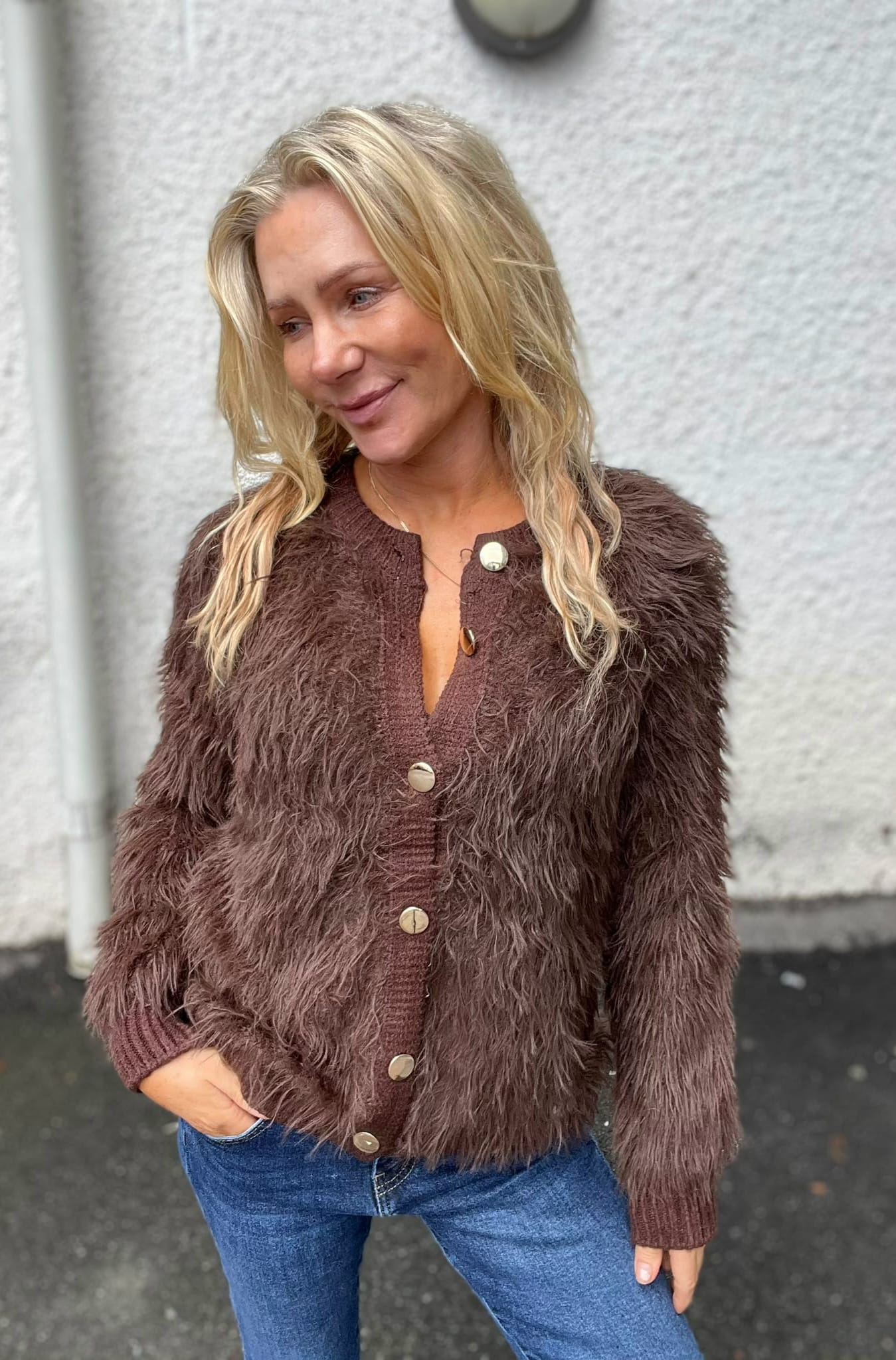 Blond kvinna står utomhus mot en ljus vägg och ler svagt medan hon tittar åt sidan. Hon bär en brun, lurvig kofta med stora knappar och blå jeans, med ena handen i fickan.