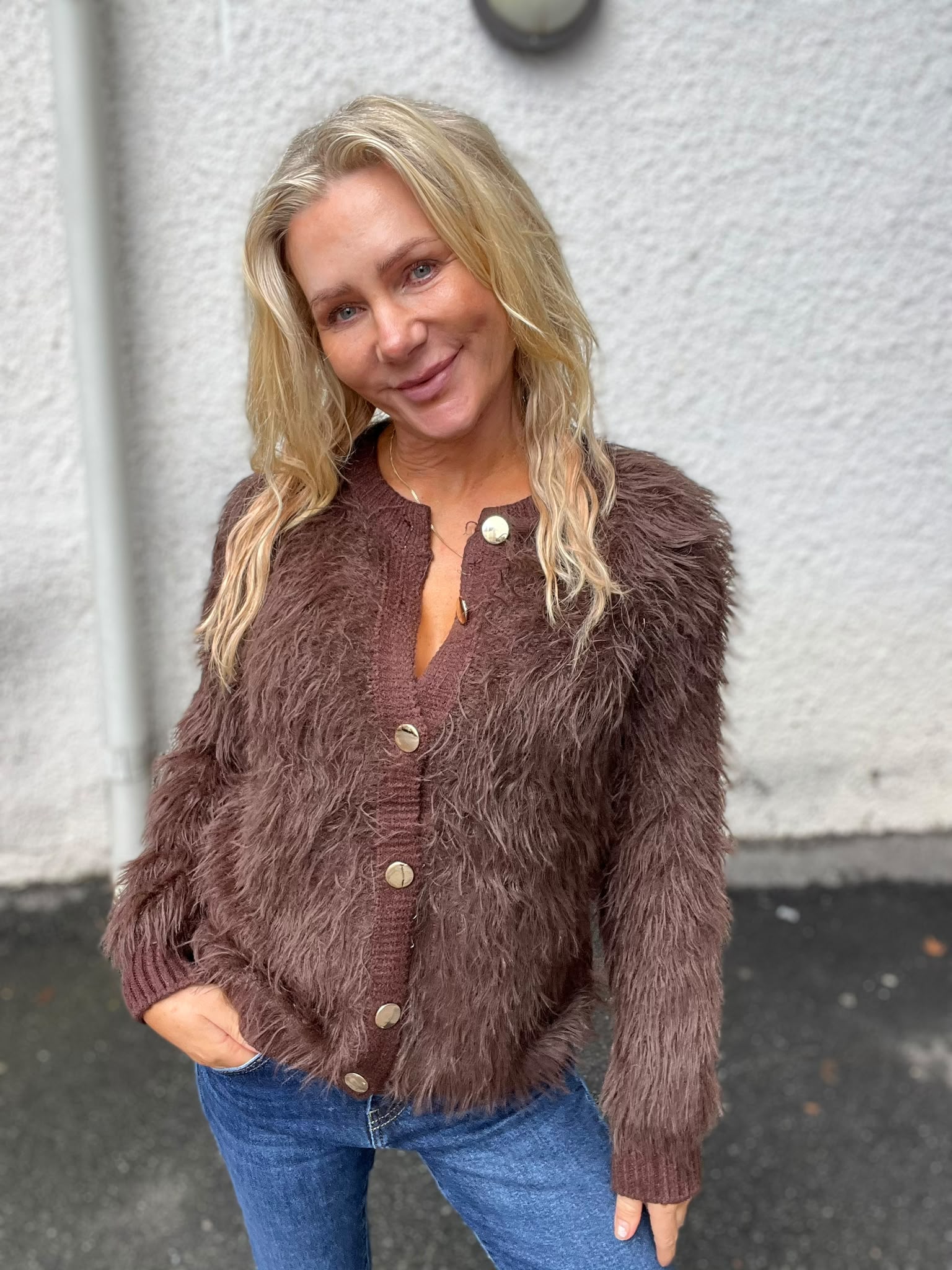 Leende blond kvinna poserar utomhus med ena handen i jeansfickan och huvudet lätt lutat. Hon bär en brun, fluffig kofta med stora knappar och halsband, framför en ljus, grovputsad vägg.