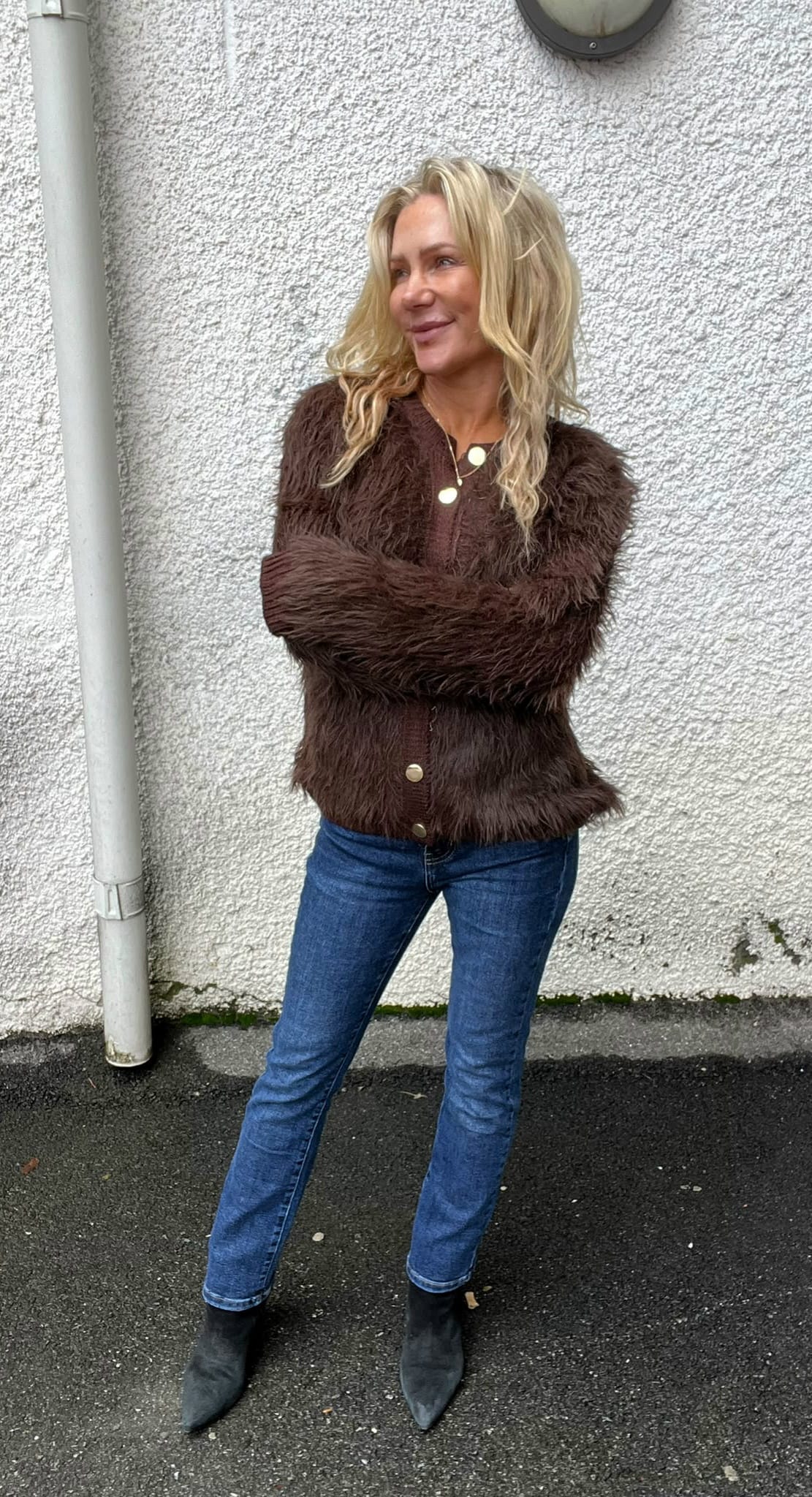 Kvinna med långt blont hår står med armarna i kors och ler, vänd åt sidan. Hon bär brun, fluffig kofta med knappar, blå jeans och svarta boots, framför en vit grovputsad vägg utomhus.