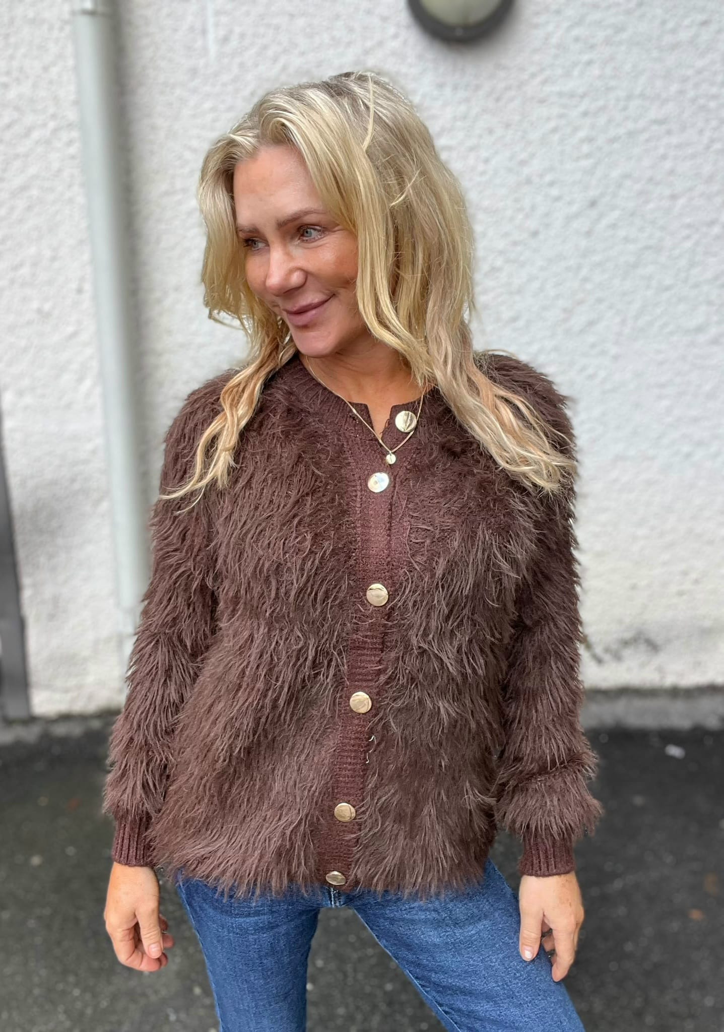 Blond kvinna står utomhus och ler medan hon tittar åt sidan. Hon bär en brun, lurvig kofta med stora knappar över en brun topp, halsband med rund berlock och blå jeans, mot en ljus vägg.