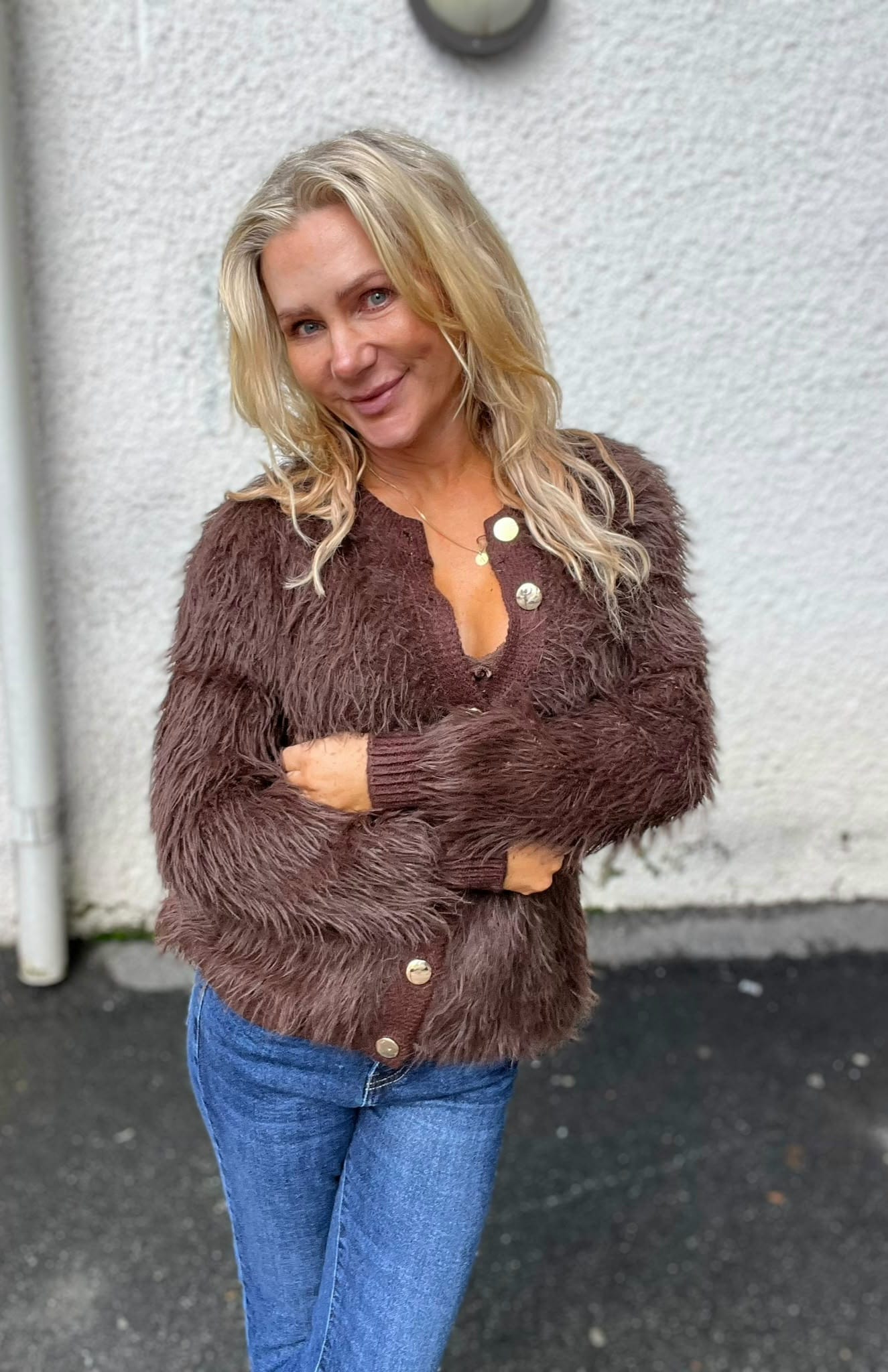 Blond kvinna står utomhus med armarna i kors och ler mot kameran. Hon bär en brun, lurvig kofta med stora knappar och blå jeans, framför en ljus, grov putsad vägg på asfalt.