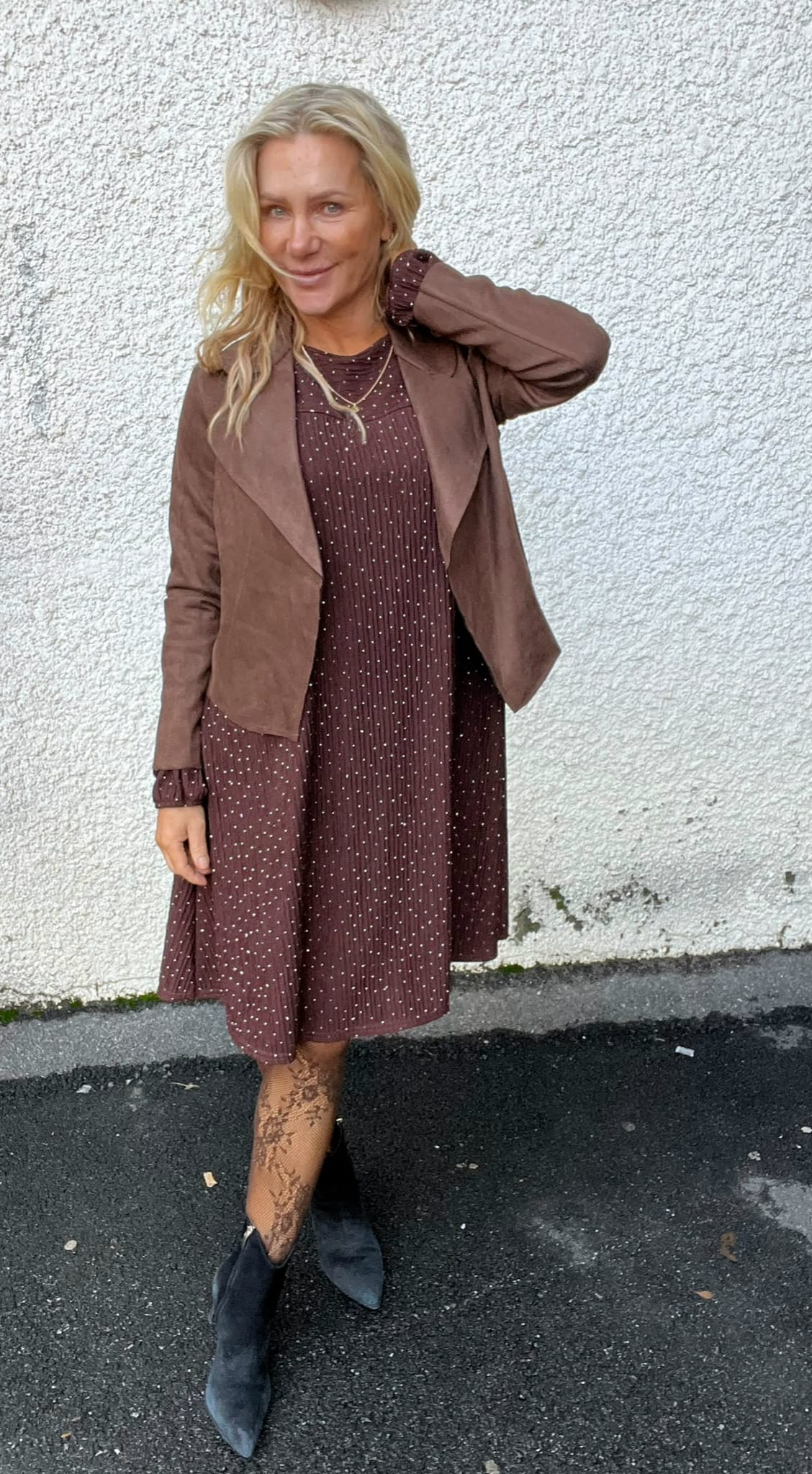 Blond kvinna poserar utomhus mot en vit putsad vägg och ler, med ena handen bakom nacken. Hon bär brun prickig klänning, kort brun jacka, mönstrade strumpbyxor och svarta boots på asfalt.