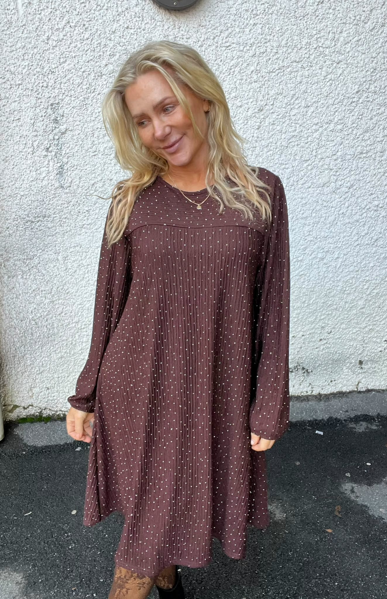 Blond kvinna står avslappnat och ler lätt medan hon tittar åt sidan, klädd i en långärmad brunprickig klänning och svarta boots. Hon står utomhus mot en vit grov vägg på mörk asfalt.