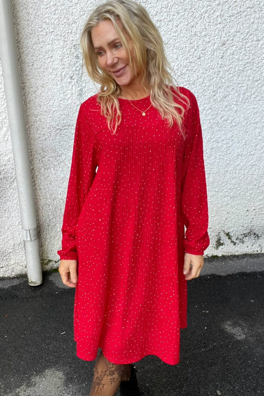Blond kvinna står utomhus och ler medan hon tittar åt sidan, klädd i röd prickig klänning med lång ärm. Hon bär halsband, mönstrade strumpbyxor och svarta skor framför en vit putsad vägg.