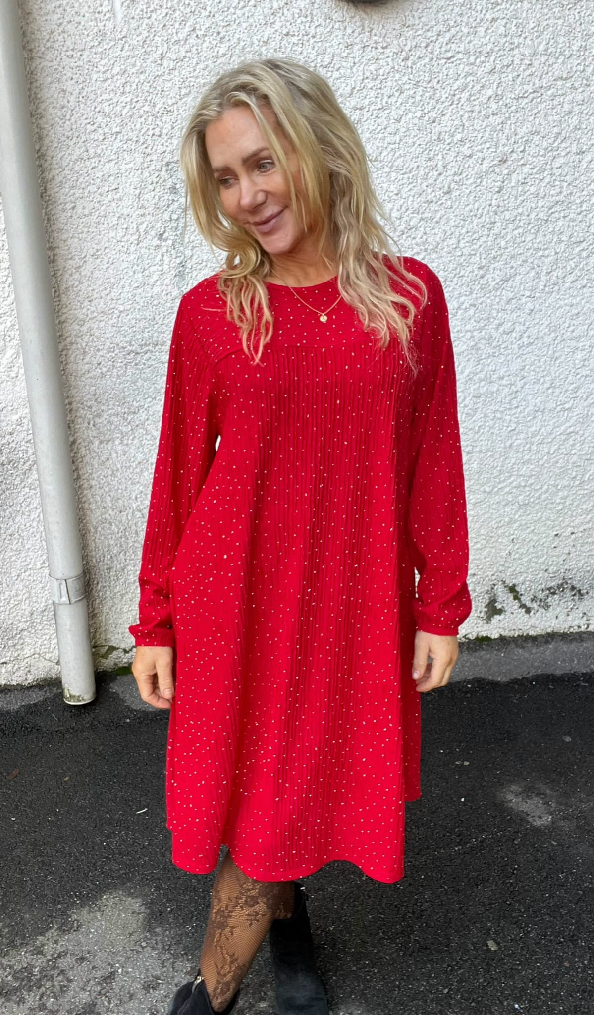 Blond kvinna står utomhus och ler medan hon tittar åt sidan, klädd i röd prickig klänning med lång ärm. Hon bär halsband, mönstrade strumpbyxor och svarta skor framför en vit putsad vägg.