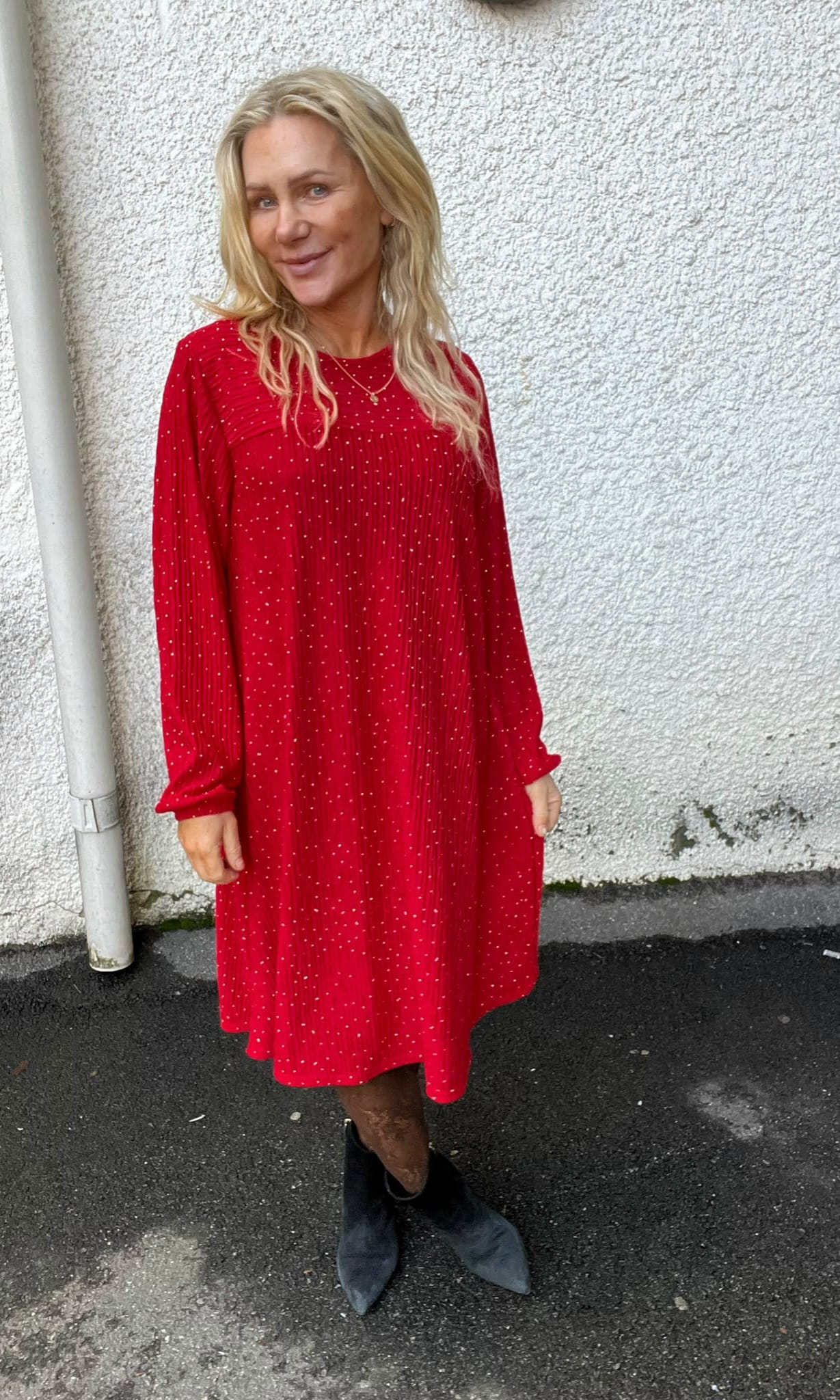 Blond kvinna står och ler lätt mot kameran framför en vit, grovputsad vägg utomhus. Hon bär en röd prickig långärmad klänning och svarta ankelboots, på asfalt bredvid ett stuprör.
