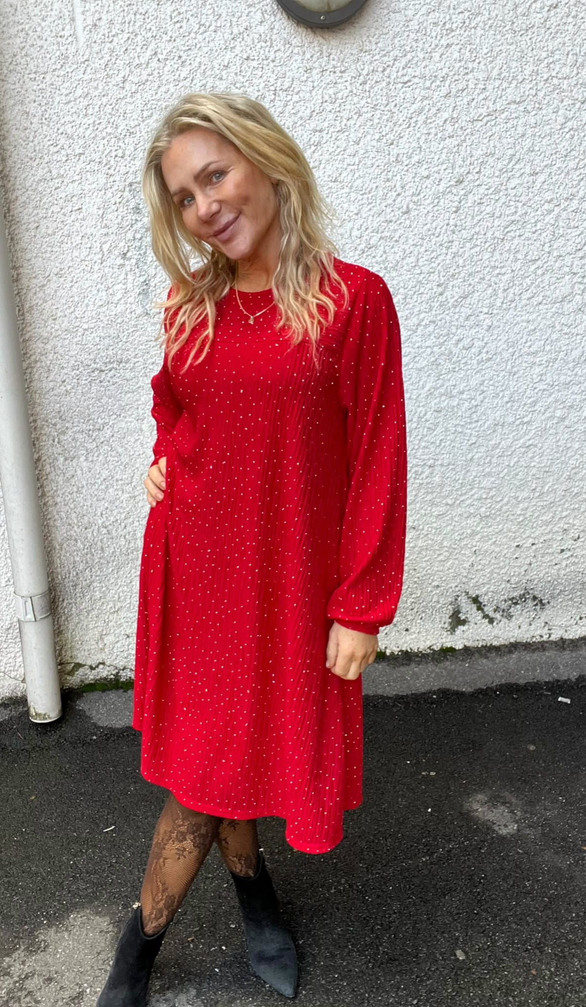 Blond kvinna står och ler lätt med handen i fickan, klädd i röd prickig långärmad klänning och svarta boots. Hon poserar utomhus mot en grov vit vägg vid en asfaltgång.