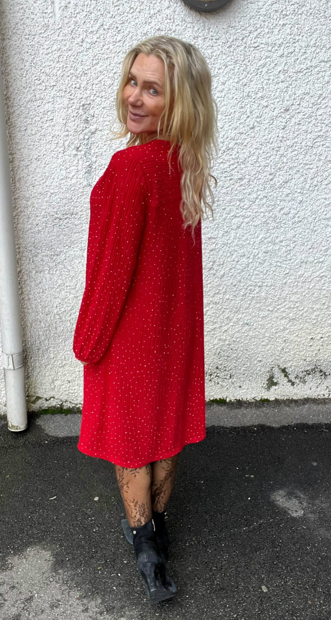 Blond kvinna står med ryggen mot kameran och vänder huvudet för att le, klädd i röd prickig klänning med långa ärmar, mönstrade strumpbyxor och svarta boots, framför en vit putsad vägg utomhus.