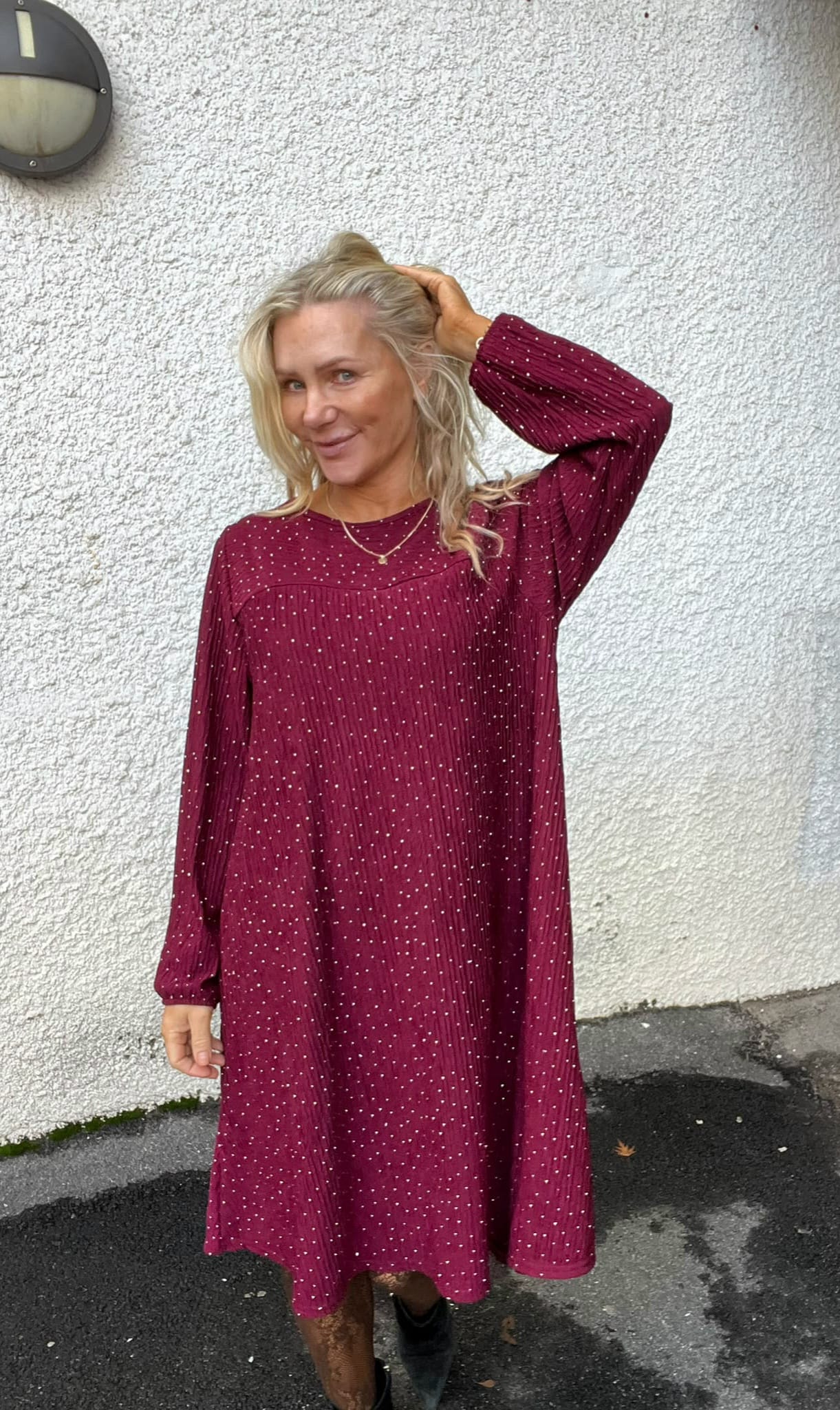 Blond kvinna står utomhus mot en vit putsad vägg och ler mot kameran, med ena handen i håret. Hon bär en vinröd, prickig långärmad klänning och svarta boots; vägglampa syns ovanför.
