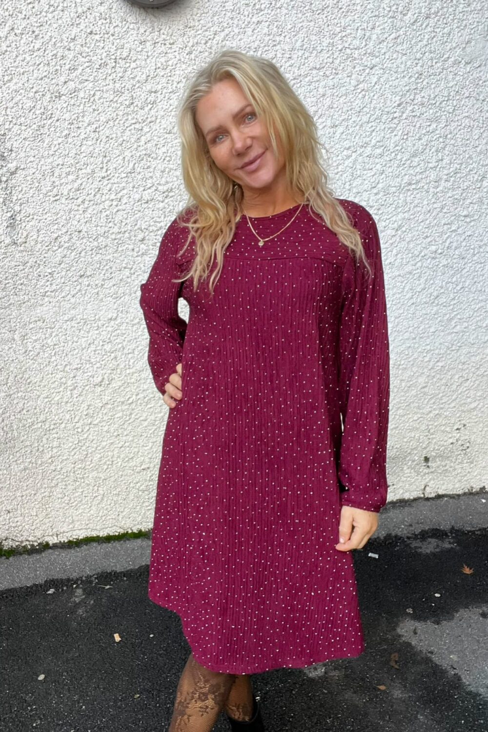 Blond kvinna står och ler mot kameran med ena handen på höften, klädd i vinröd klänning med små vita prickar och tunna halsband. Hon står utomhus framför en vit putsad vägg på asfalt.