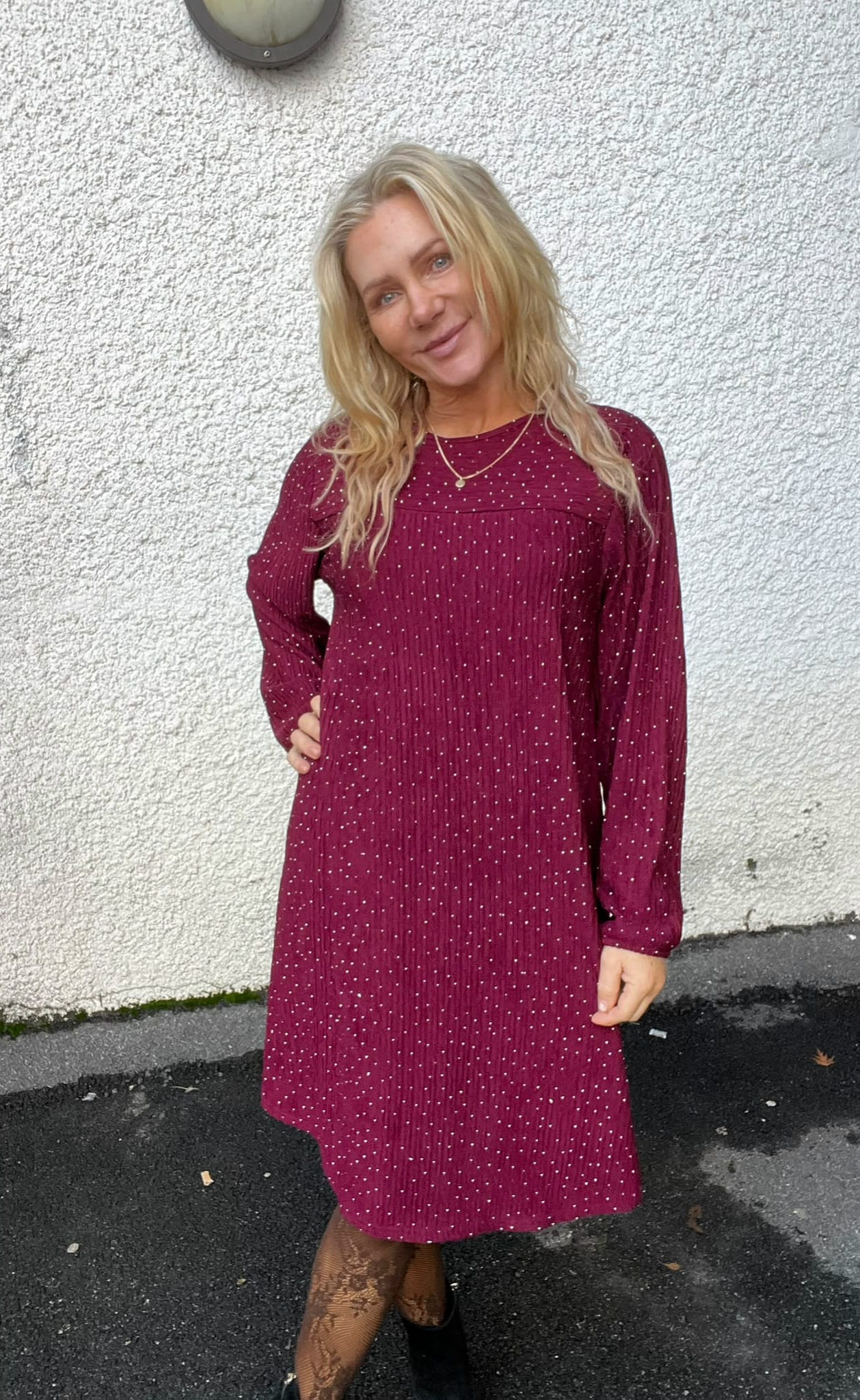 Blond kvinna står och ler mot kameran med ena handen på höften, klädd i vinröd klänning med små vita prickar och tunna halsband. Hon står utomhus framför en vit putsad vägg på asfalt.
