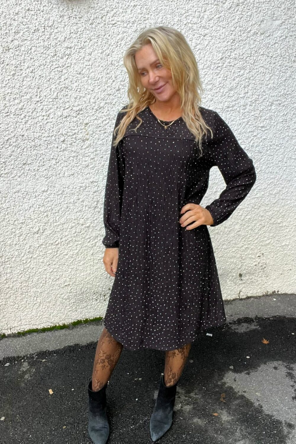 Blond kvinna står med ena handen på höften och tittar åt sidan, iklädd svart långärmad prickig klänning, mönstrade strumpbyxor och svarta spetsiga boots, framför en vit putsad vägg utomhus på asfalt.