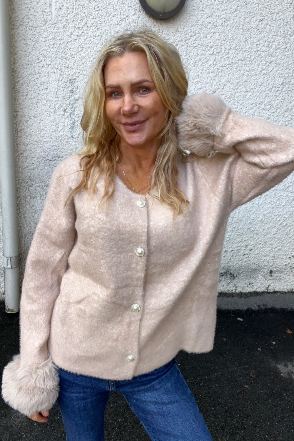 Blond kvinna står utomhus och ler mot kameran med ena handen bakom huvudet. Hon bär en ljusrosa, fluffig kofta med stora knappar och jeans, framför en vit, grovputsad vägg med rör och vägglampa.